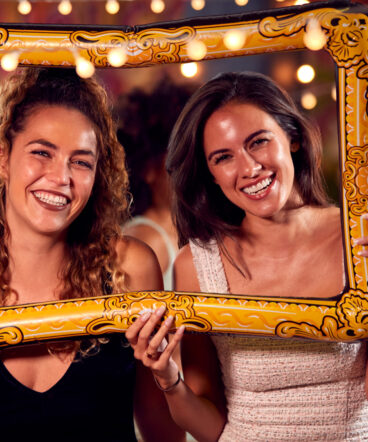 Female Friends Having Fun Posing With Photo Booth Photo Frame At Party In Bar